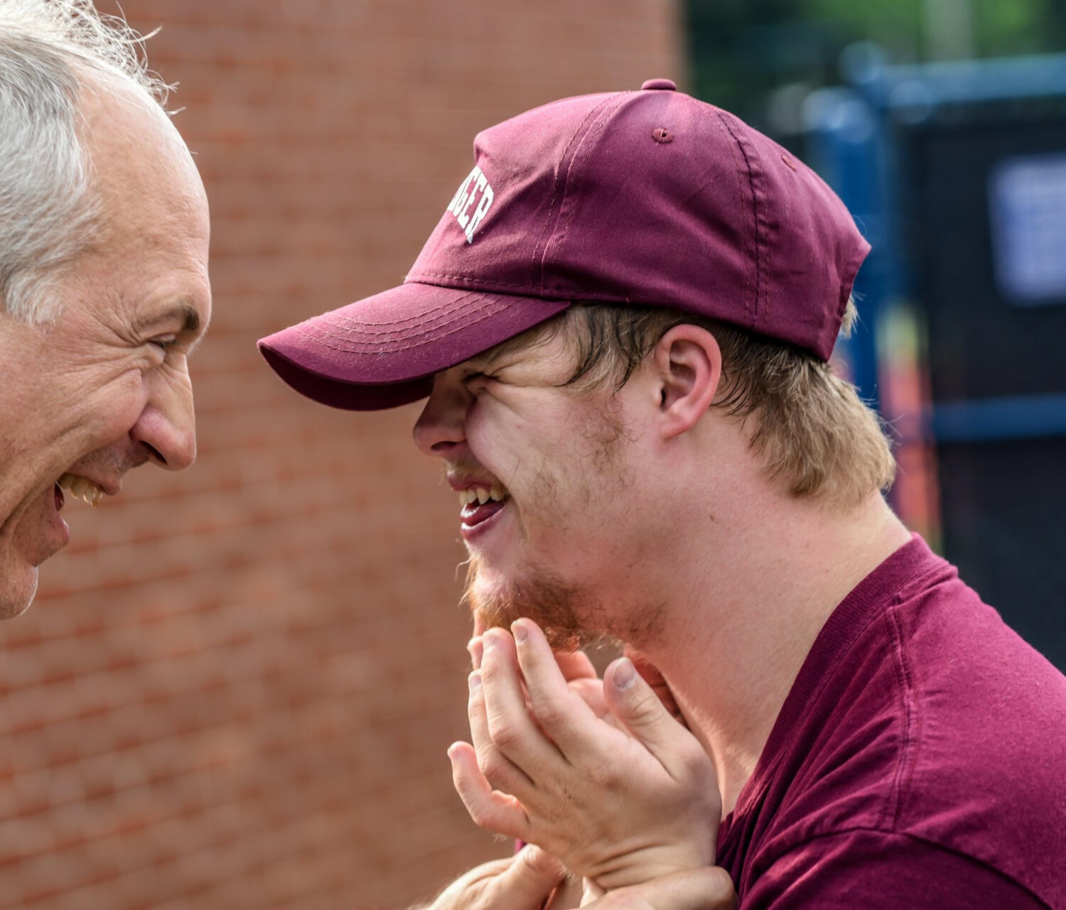 An older man with white hair, looking and smiling at a younger man with light brown hair and facial hair, wearing a red hat, smiling back at the older man.